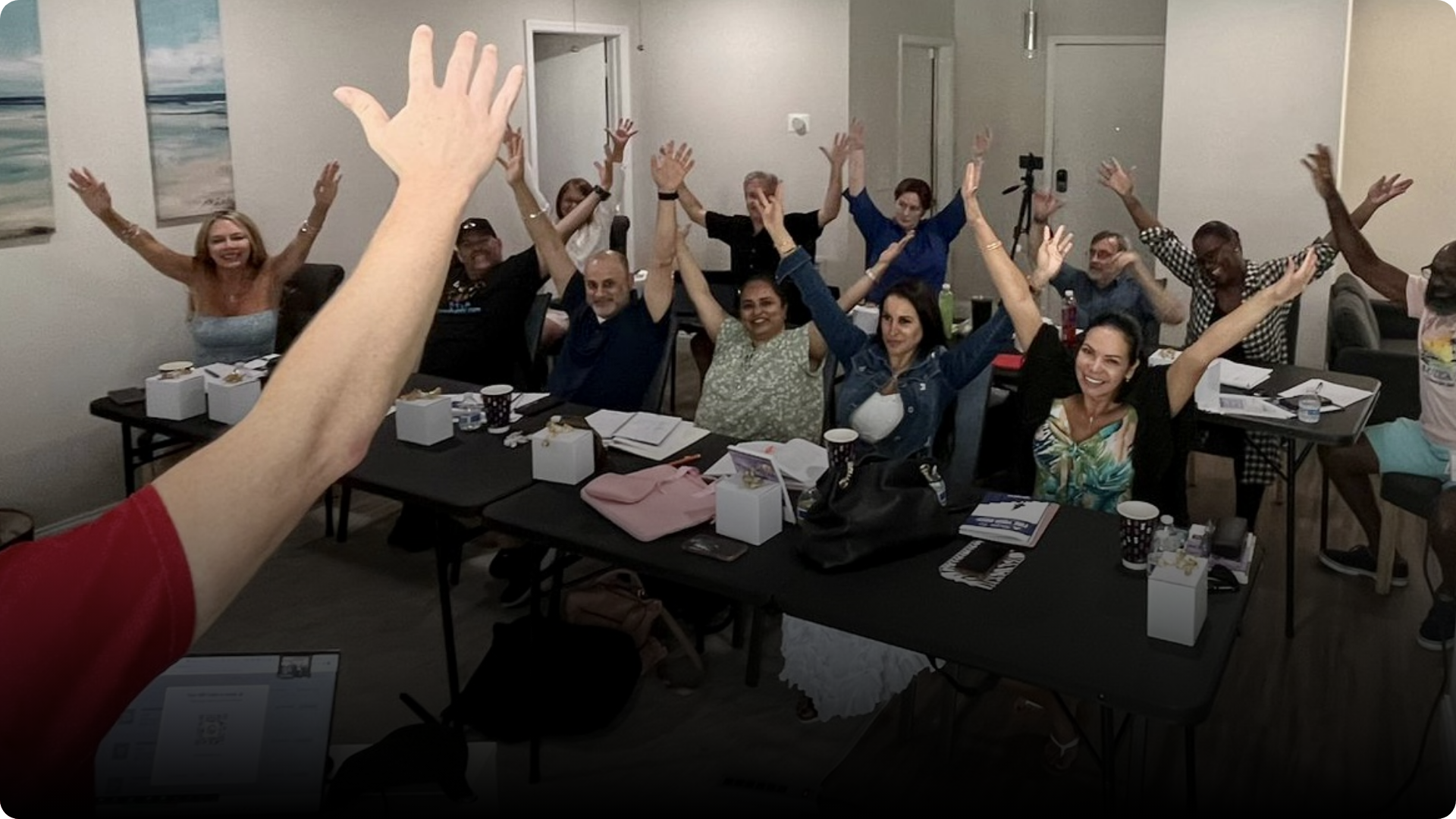 Group of enthusiastic participants at Hugh Zaretsky's real estate boot camp workshop, raising hands in celebration, with tables featuring notebooks and materials, led by a presenter in a red shirt.