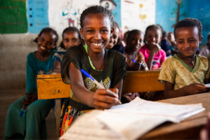 Smiling African girl writing in a notebook surrounded by classmates in a classroom, highlighting educational initiatives supported by Hugh Zaretsky's charitable work for community impact.