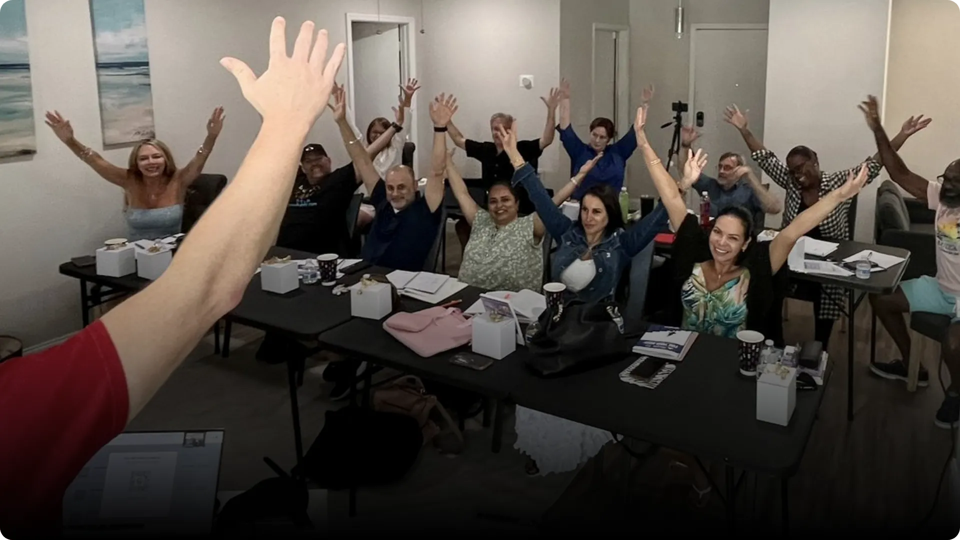 Group of enthusiastic participants at Hugh Zaretsky's real estate boot camp workshop, raising hands in celebration, with tables featuring notebooks and materials, led by a presenter in a red shirt.